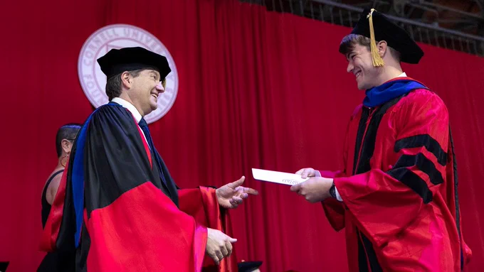 Two individuals in academic regalia are on a stage with a red backdrop and a Cornell University emblem. One person is handing a certificate to the other, who is smiling.