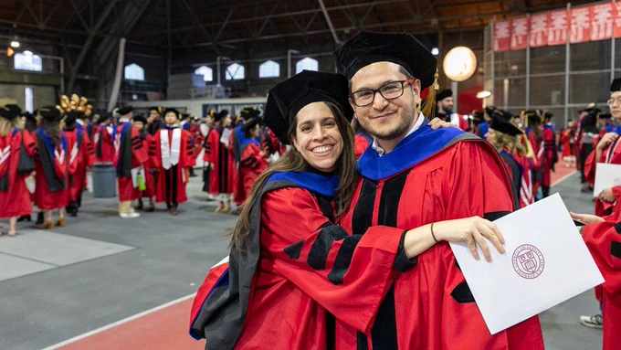 Two Cornell graduates in red and black gowns celebrate indoors. One person holds a diploma with a seal. A large group in similar attire is seen in the background.