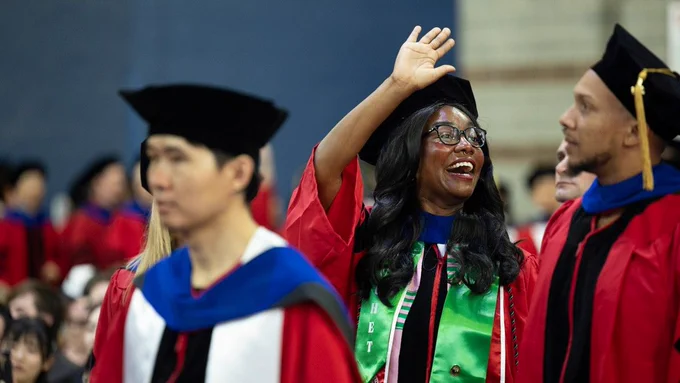 A group of Cornell graduates in red academic robes and black caps are celebrating. One person is smiling and waving. The background shows other graduates and an audience.