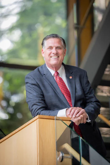 A white man leans against a podium, smiling and posing for a picture. 