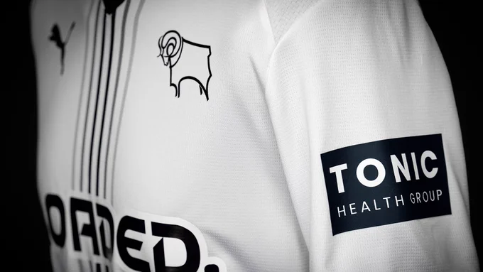 An image from the side of Derby County's new white 2026/27 PUMA home shirt - with six greyscale pinstripes down the centre, the Loaded sponsor logo across the front and the Tonic Health Group logo on the left sleeve. The shirt also includes a black V-neck collar and PUMA and Derby County logos. 
