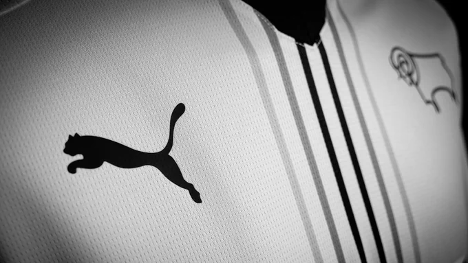 An close-up image of the front of Derby County's new white 2026/27 PUMA home shirt - with six greyscale pinstripes down the centre. 
The image focused on the PUMA logo, with the Derby County ram crest out of focus on the opposite side. 