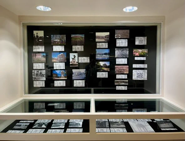 A display case featuring various photographs and printed documents. The images are arranged on a black background within a well-lit frame. There are two levels of the display, with additional photographs and documents at the lower level.