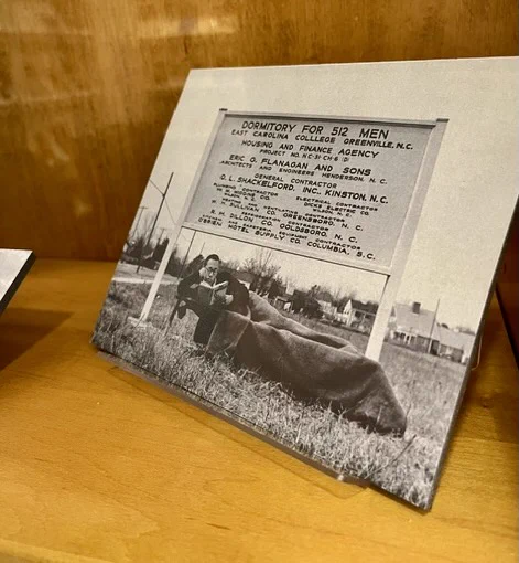 A vintage photograph displayed in a frame shows a person laying at a sign for a dormitory project in Greenville, North Carolina. 