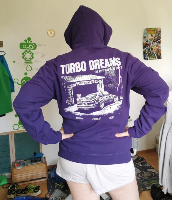 Person steht mit dem Rücken zur Kamera in einem hellen Zimmer und trägt einen lila Hoodie mit großer weißer Rücken-Grafik. Aufdruck mit dem Schriftzug „TURBO DREAMS“ und einer Retro-Auto-/Tuning-Szene vor einer Garage im Streetwear-Stil. Kapuze aufgesetzt, Hände in die Hüften gestemmt. Dazu helle kurze Shorts. Der Pullover wirkt lässig, modern und von Motorsport- bzw. JDM-Kultur inspiriert.
