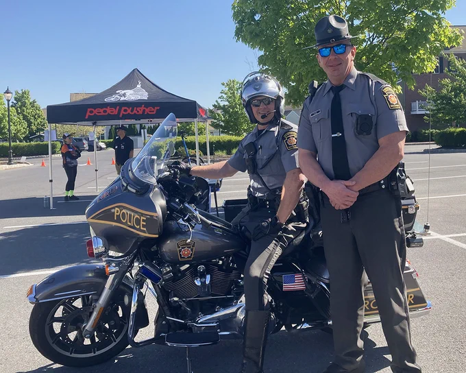 Outdoors on a sunny day, two Troopers pose in a parking lot; one sits on a marked police motorcycle