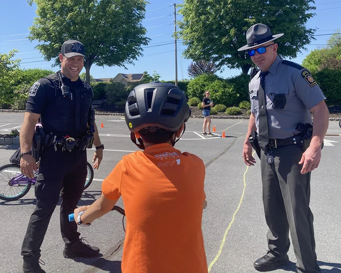 Outdoors on a sunny day, a young bicyclist interacts with a Cumberland County Sheriff’s Office officer and a Trooper