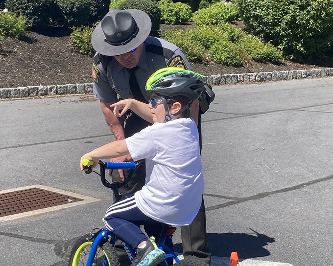 Outdoors on a sunny day, a young bicyclist gestures while talking with a Trooper