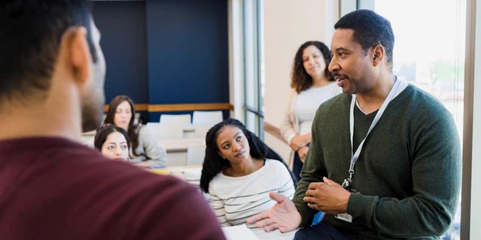 A teacher in front of students in a classroom with a few of his colleagues looking on