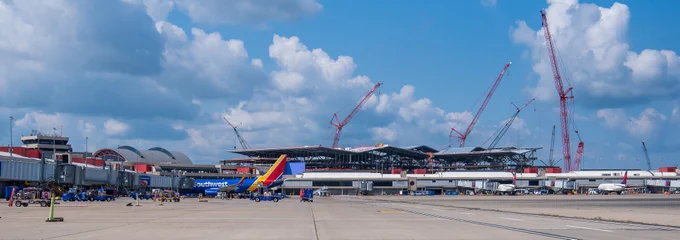 Wide view of an airport terminal under construction with multiple cranes and parked airplanes on a sunny day.