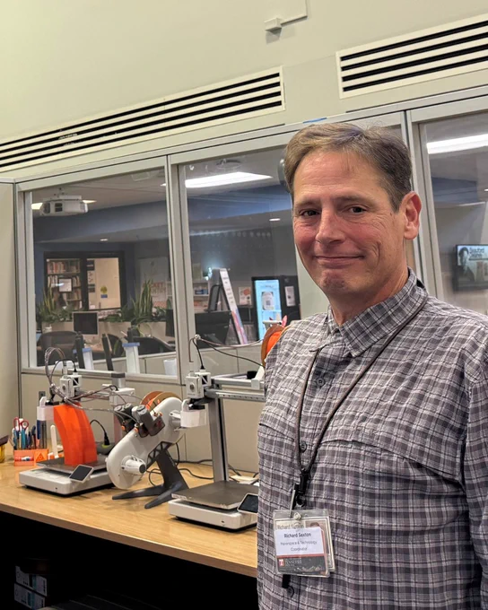 A staff member stands in a makerspace next to several 3D printers and equipment on a workbench.