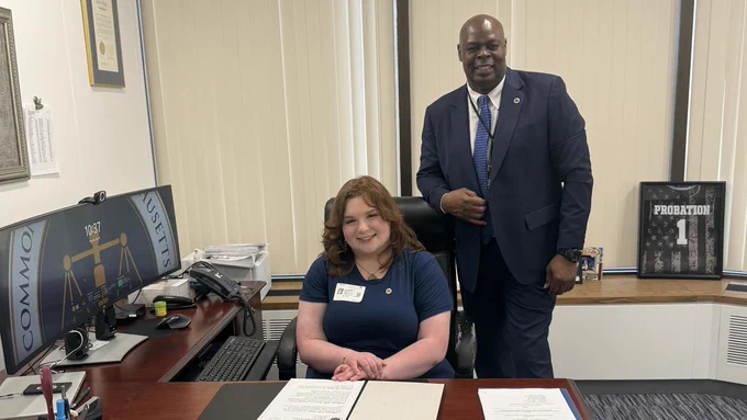 A student participant in the Massachusetts Probation Service “Co-Commissioner for the Day” program sits at a desk in a probation office, smiling toward the camera, while Probation Commissioner Edward Dolan stands beside her. Computer monitors and office materials are visible in the workspace.