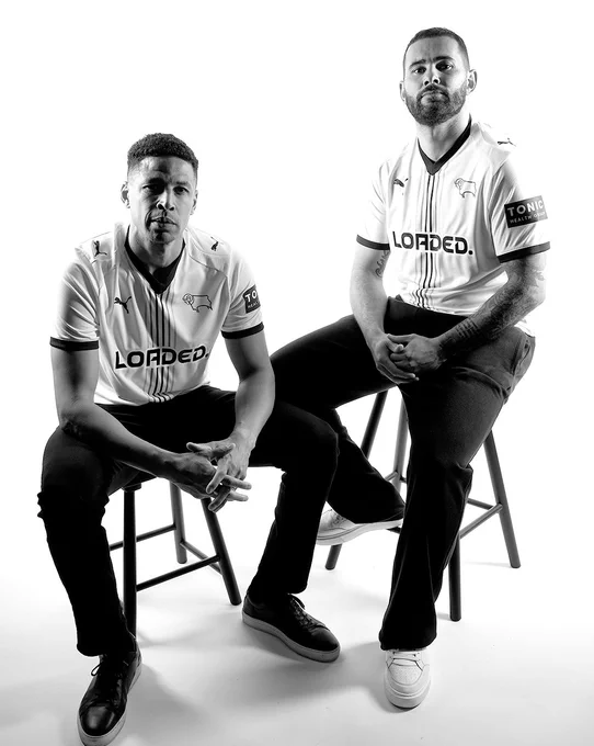 Former Rams players Curtis Davies and Bradley Johnson modelling Derby County's new 2026/27 PUMA home kit. The duo are sat on black wooden stools in a photography studio with a white background.