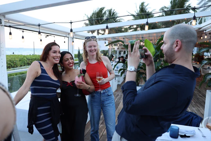 One coworker takes a picture of three others. They are all smiles at a beach front property.