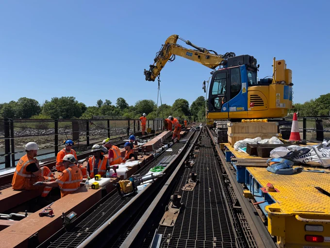 Railway engineers on track working on a bridge, with a large yellow machine with an arm lifting a large wooden block into place.