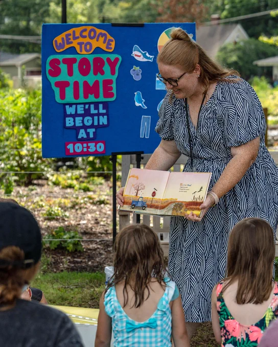 Woman in patterned dress reading a children's book aloud to kids during an outdoor story time. A board behind her reads Welcome to Story Time. We'll begin at 10:30