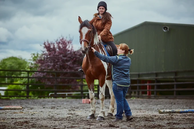 Woman sitting on a horse in a muddy field, while someone on the ground stands and attends to its reins.