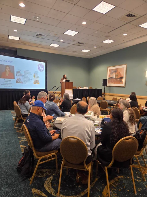A close up photo of the conference room with a slideshow at the front which includes the Hempstead Union Free School District logo. Tiara Adams stands behind the podium speaking to the crowd, who sits around circular tables. The chairs are brown and the floor has a patterned carpet. 