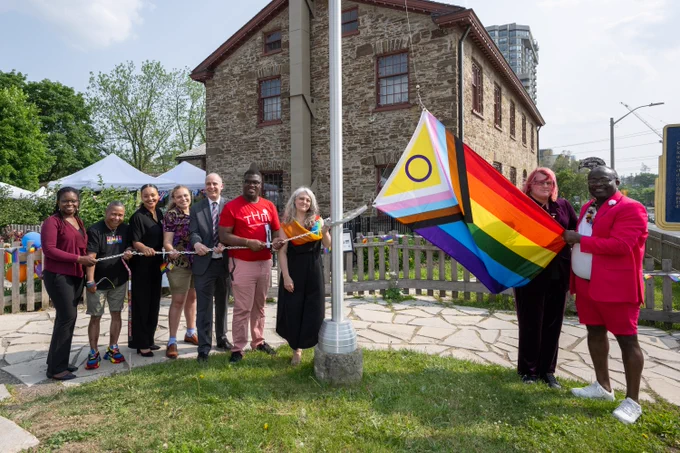 Group of diverse people raising a Progress Pride flag outside a historic stone building on a sunny day.