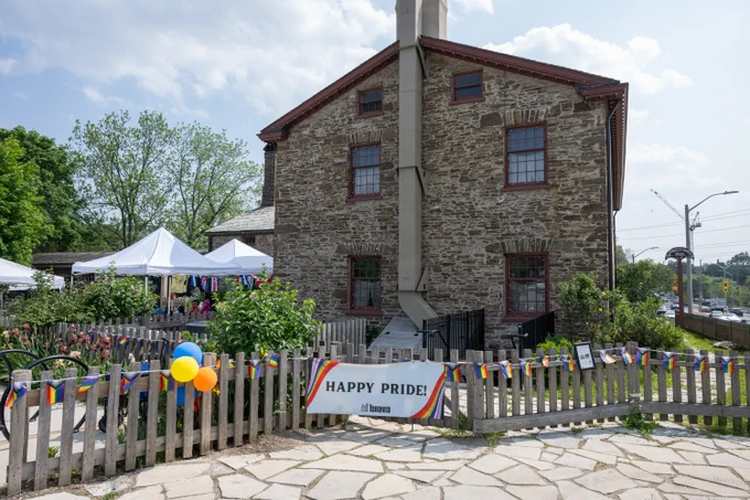 Stone building with a wooden fence decorated with colorful Pride flags and balloons, featuring a Happy Pride sign in front.