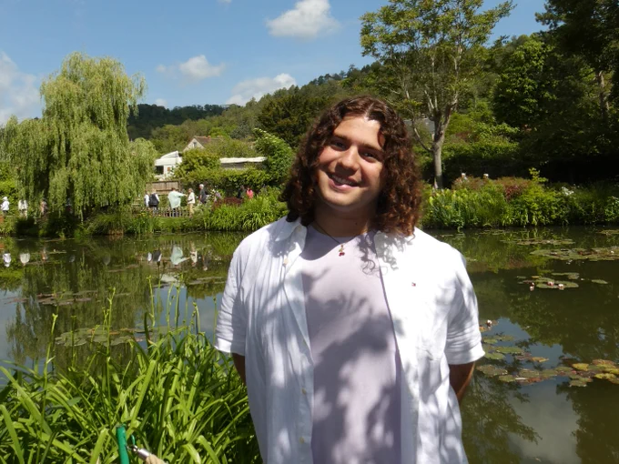 A person with curly hair standing in front of a pond filled with water lilies and surrounded by lush greenery. There are trees and a clear blue sky in the background.