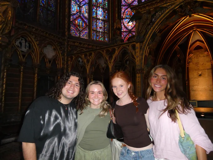 A group of four people stands inside a richly decorated room with ornate architecture and vibrant stained-glass windows.