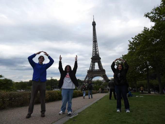 Three people are posing playfully in front of the Eiffel Tower, forming the letters "O," "H," and "I" with their arms. They are standing on a path surrounded by grass and trees, with a cloudy sky overhead.