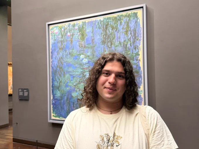 A person with long, curly hair stands in front of a framed painting in a museum. The artwork is one of Claude Monet's Water Lilies series.