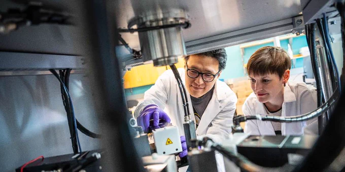 Two scientists in lab coats and gloves examine the contents of a metal chamber equipped with high-tech components. 