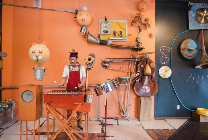 Man playing a xylophone in front of an orange wall with whimsical musical contraptions and sculptures.