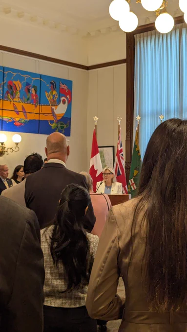 Attendees stand during the National AccessAbility Week MPPs Breakfast Reception at the Ontario Legislature while a speaker addresses the room from a podium. Canadian, Ontario, and Saskatchewan flags are displayed behind the speaker, with artwork and historic architectural details visible throughout the chamber. 