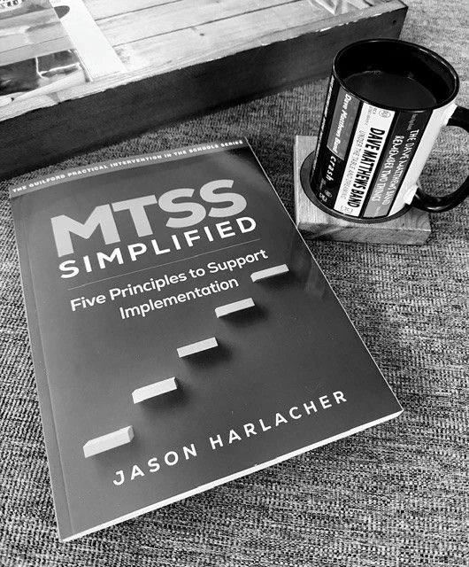 Black and white photo of a book titled “MTSS Simplified” sitting on a table next to a coffee cup. 