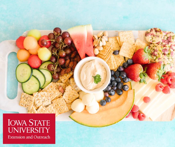 Colorful fruit and cheese platter with crackers, hummus, and nuts on a wooden board against a blue background.