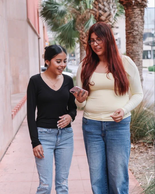 Two people walking along a sidewalk. One person is showing the other their phone.