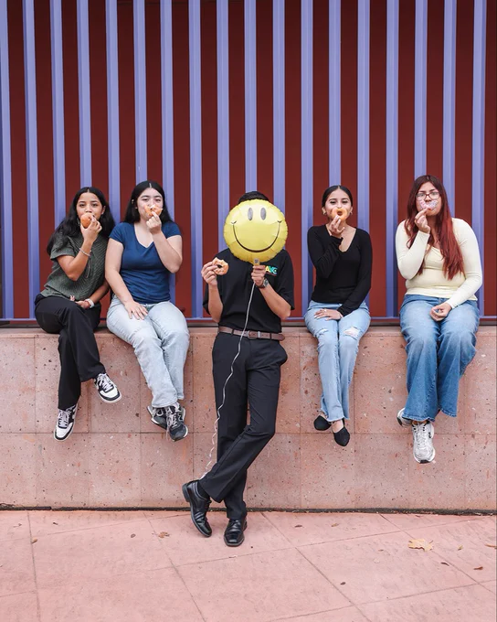 4 people sitting while holding donuts up to their face while a person in the center is holding a balloon with a happy face.