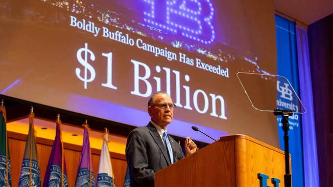 President Tripathi, a man in glasses & a suit, standing behind a podium with flags on the left. The screen behind him reads, \"Boldly Buffalo Campaign Has Exceeded $1 Billion\". A logo for UB university can also be seen to the right.
