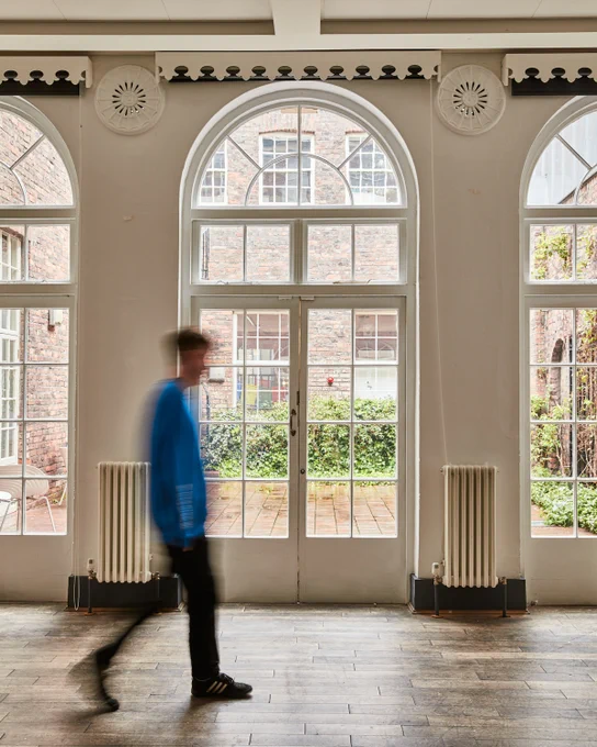 A person in motion walks past large arched windows with white frames. The windows overlook a brick-walled courtyard with greenery. The setting features a wooden floor and decorative heating elements beneath the windows.