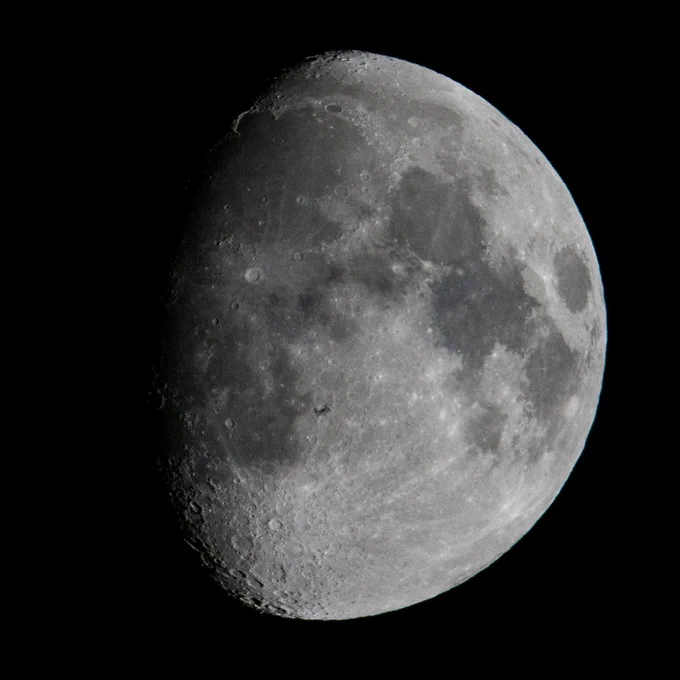 A close-up photo of the Moon, which is about three-quarters full; its left side is in shadow. Its gray seas and circular impact craters are clearly visible. Near the center of the image, the International Space Station can just barely be seen passing in front of the Moon. Credit: NASA/Joel Kowsky