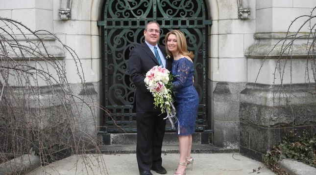 A couple is standing arm in arm in front of an ornate wrought iron gate set in a stone building. They are dressed formally; one holds a large bouquet of white and pink flowers.