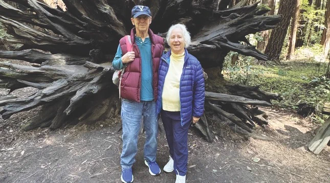 Two people stand close together in front of a large, intricate tree root in a forest setting. One wears a cap and a red vest, while the other is in a blue jacket. The scene is peaceful and surrounded by nature.