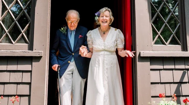 An elderly person in a suit and another in a white dress are joyfully stepping out of a building with a brown exterior and diamond-patterned windows. They are holding hands and surrounded by floral decorations.