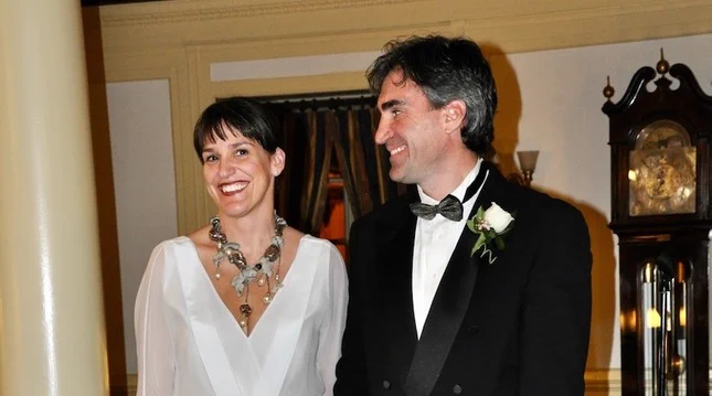 Two smiling individuals. One is wearing a white dress and the other is wearing a black tuxedo.