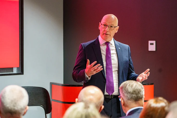 First Minister John Swinney speaks to an audience in a modern conference room.