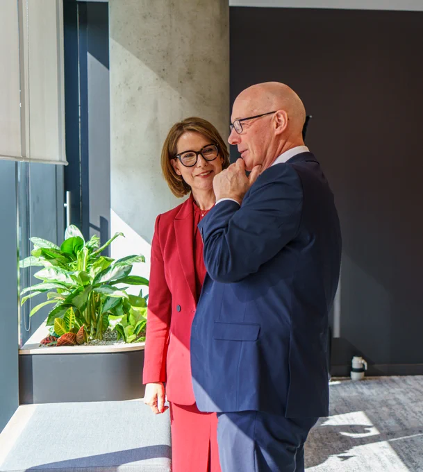 First Minister John Swinney and Hitachi Energy Country Managing Director UK and Ireland Laura Fleming are stood at a window in business attire.