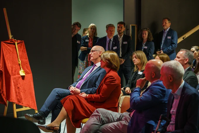 Audience, including First Minister John Swinney, seated attentively in a dimly lit room facing a covered easel.