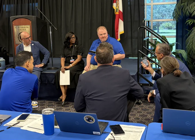 A group of individuals is seated in a discussion at an SPC meeting room. Laptops and documents are on the table. A flag and large window with a view of greenery are in the background.