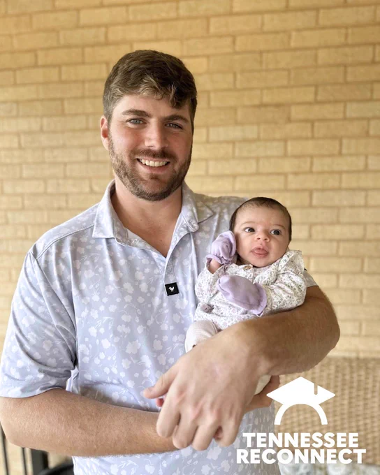 Smiling man wearing a light patterned polo shirt holding a young baby dressed in a floral outfit with purple mittens against a brick wall background.
