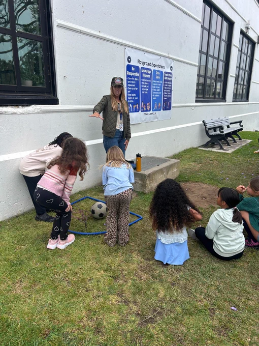 Teacher instructing a group of young children gathered on grass near a building with a soccer ball inside a blue hoop.