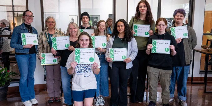 Group of 10 students standing indoors holding certificates, celebrating together in a bright room with large windows.