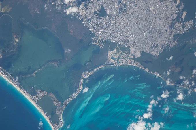 (08/30/2014) -- Cancún, Mexico. A long lens was used by astronauts aboard the International Space Station to take this image, and it highlights many natural and built features. The street pattern of Mexico’s tourist mecca, Cancún, contrasts with the waterways of the marinas that open into the bay and the lagoons. Brilliant blue water over coral reefs contrast with the dark waters of inland lagoons. The reefs are the second largest reef system on Earth, and draw tourists from all over the world. The wide, well developed beach on the gulf coast (image upper right) is the result of vigorous wave energy; the white sand makes the beach easily visible from space. But wave energy is reduced along Cancún’s protected shoreline (image center) and the beaches are thin or non-existant. Fair-weather cumulus clouds are scattered across the image top left.